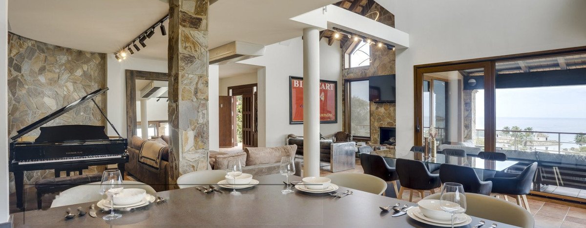 Spacious living and dining area with a grand piano, elegant dining table, and a stunning sea view through large glass doors.