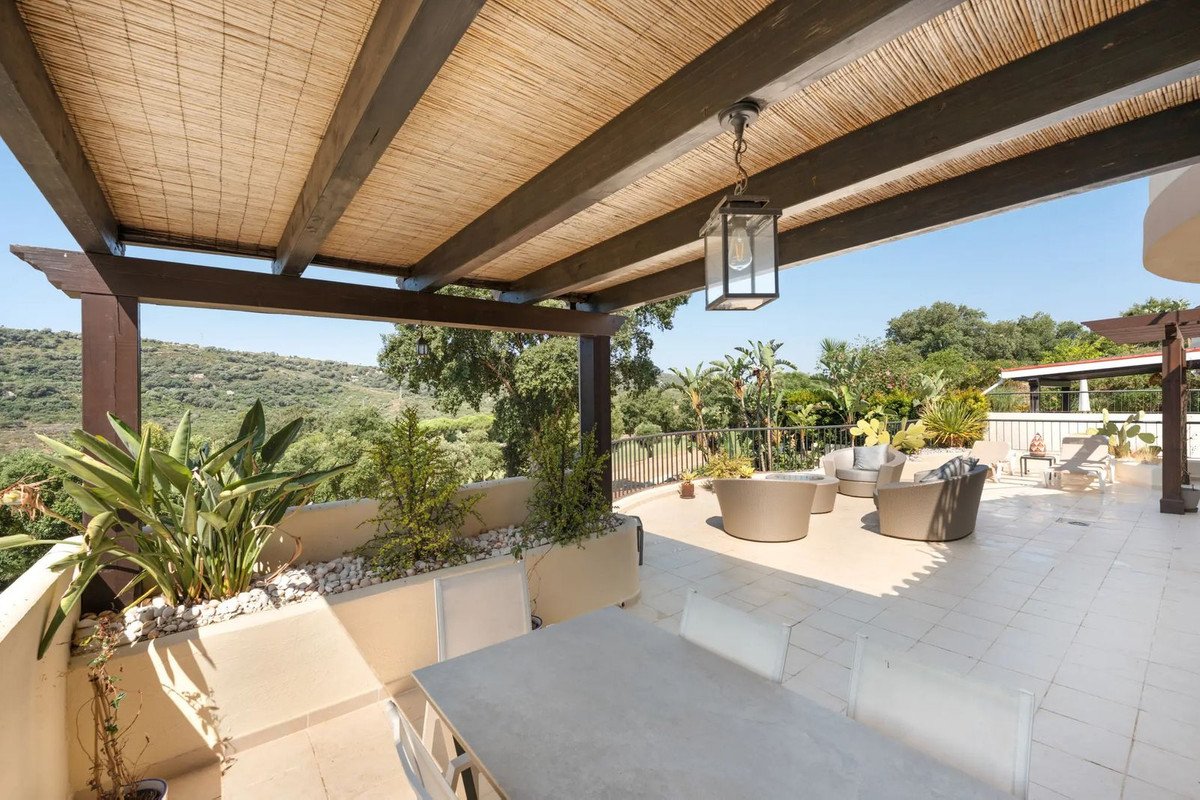 A spacious terrace with outdoor seating furniture and lush greenery overlooking a scenic landscape under a bamboo-covered pergola.
