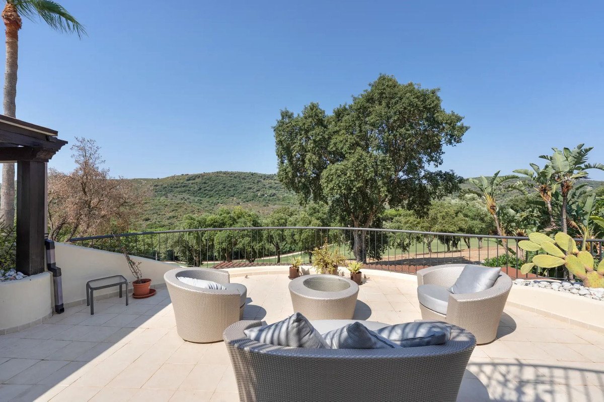 Outdoor terrace with modern rattan seating and a scenic view of rolling green hills and trees under a clear blue sky.