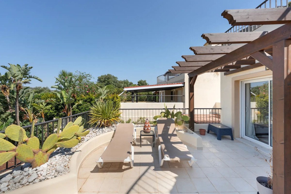 Terrace with two lounge chairs, a pergola, and lush greenery providing a scenic view under a clear blue sky.
