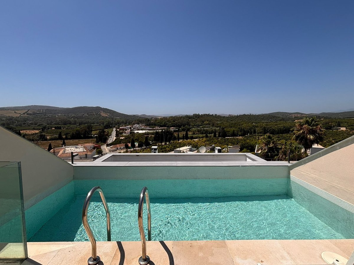 An infinity pool with metal ladder overlooks a lush green valley and distant hills under a clear blue sky.