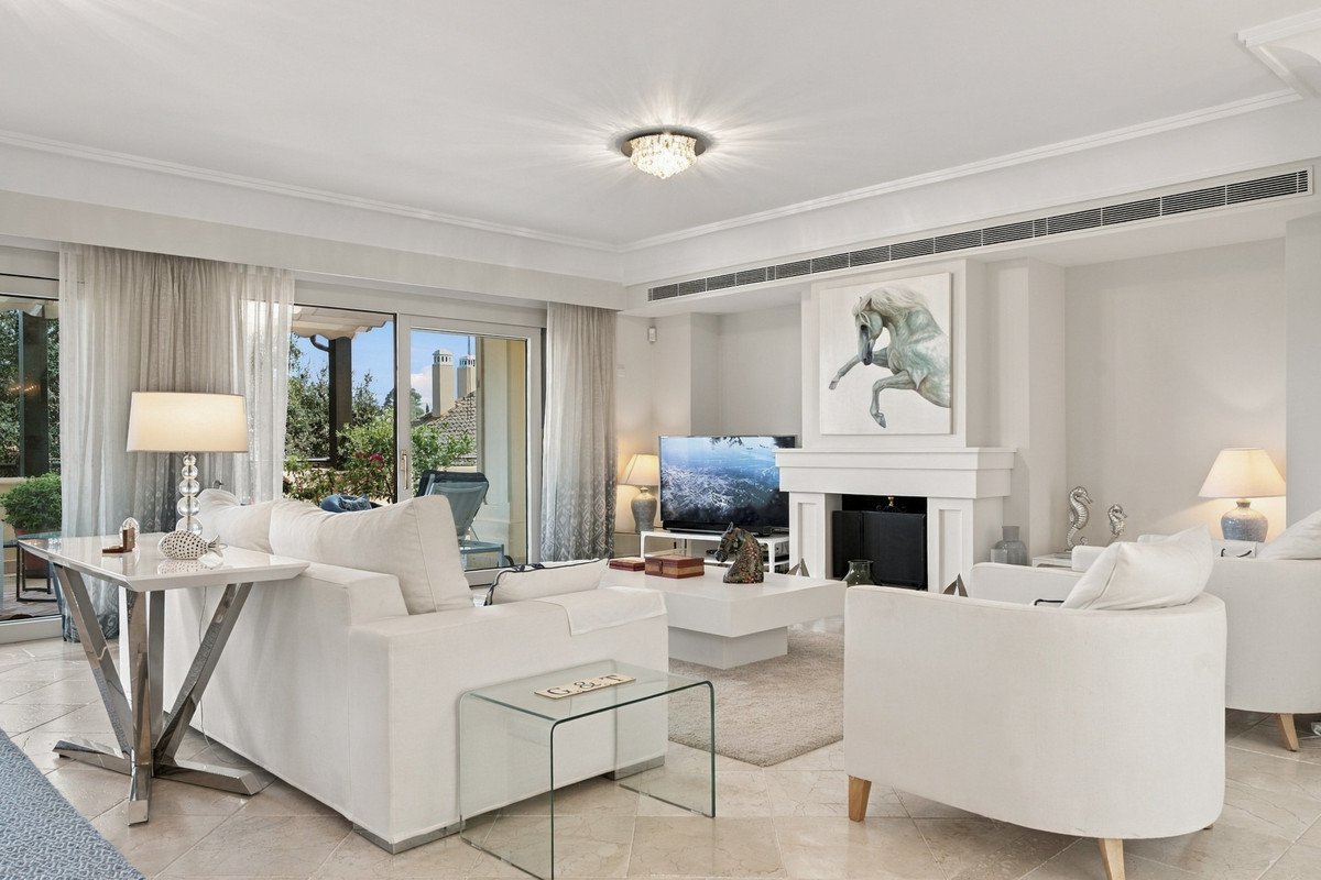 Spacious living room with white furniture, fireplace featuring horse art, and access to a terrace with greenery and distant buildings.