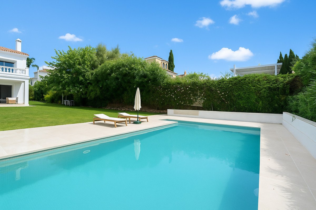 A rectangular swimming pool with turquoise water, surrounded by a paved area with lounge chairs and an umbrella, overlooking a lush garden.
