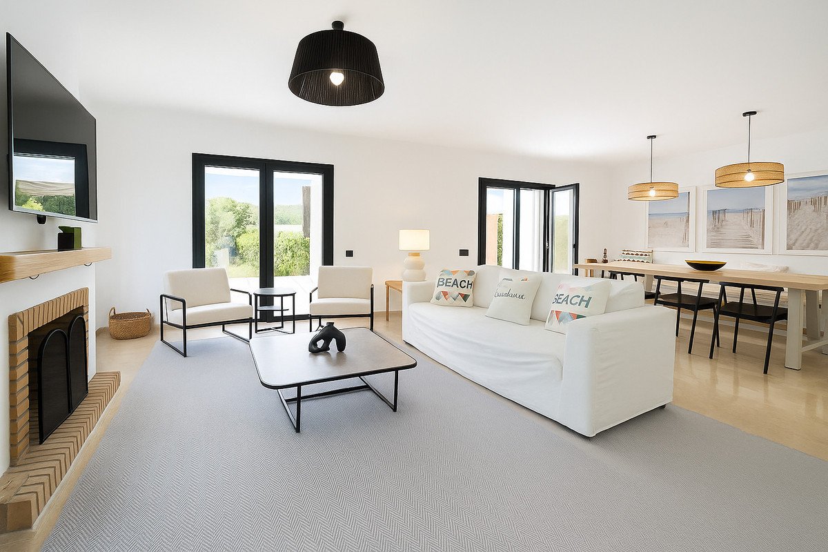 Modern living room with fireplace, white sofa, chairs, and a large dining table, featuring garden views through glass doors.