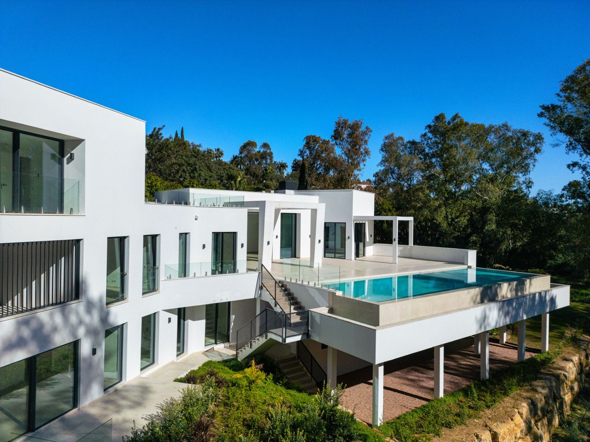 A modern white villa with an infinity pool overlooking a lush green landscape under a clear blue sky.