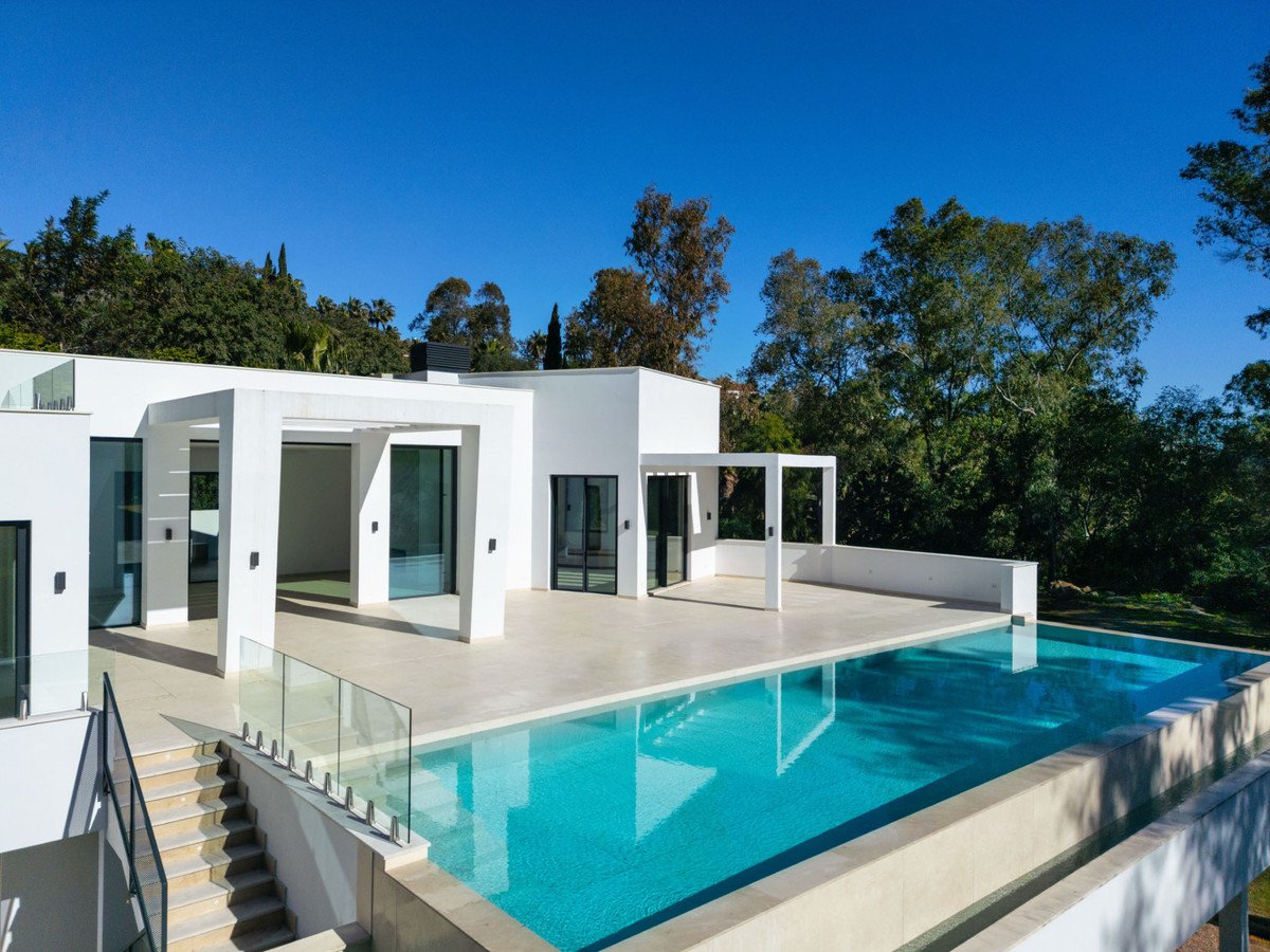Modern white villa with a large infinity pool and tiled terrace, surrounded by lush green trees under a clear blue sky.