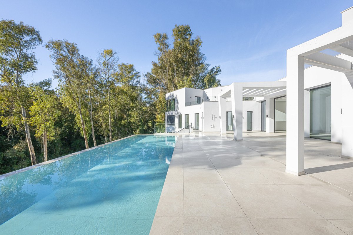 Modern white villa with an infinity pool and spacious outdoor patio surrounded by trees.