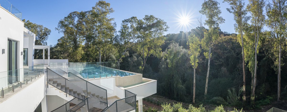 Modern white villa with an infinity pool and glass railings, surrounded by lush trees under a sunny sky.