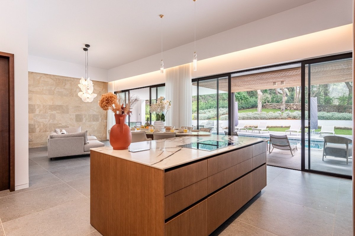 Modern kitchen with island and integrated stovetop, opening to a living area and a pool with garden view.