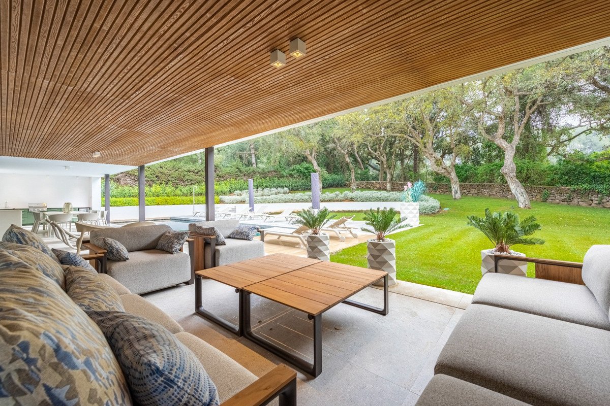Spacious covered terrace with comfortable seating, wooden table, overlooking a lush garden and swimming pool.