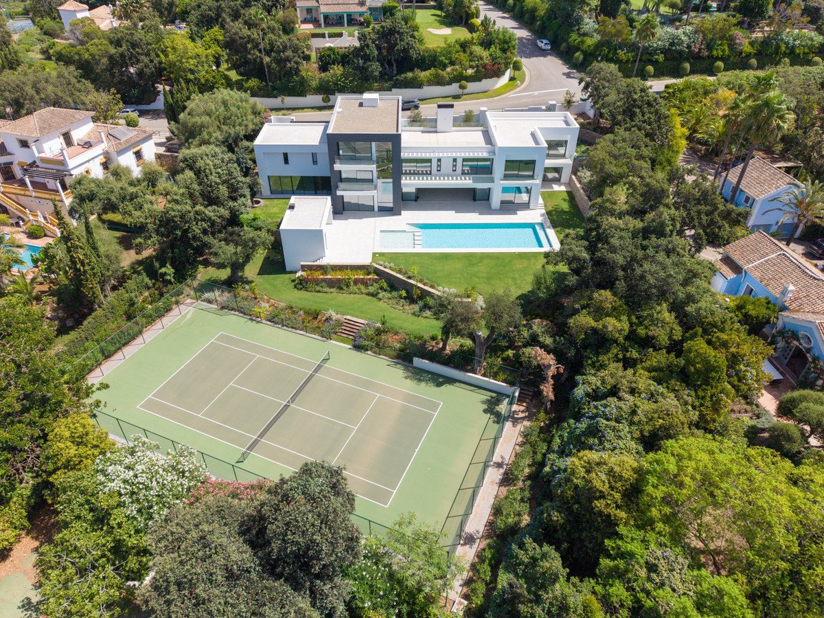 Aerial view of a modern villa with a swimming pool and a private tennis court surrounded by lush greenery.