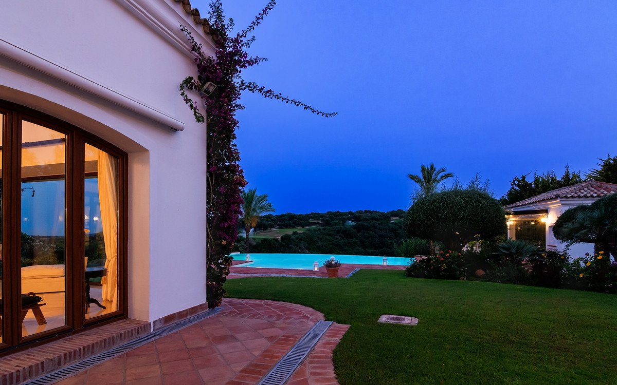 View of a illuminated infinity pool and garden with a terrace and large glass doors, at dusk.