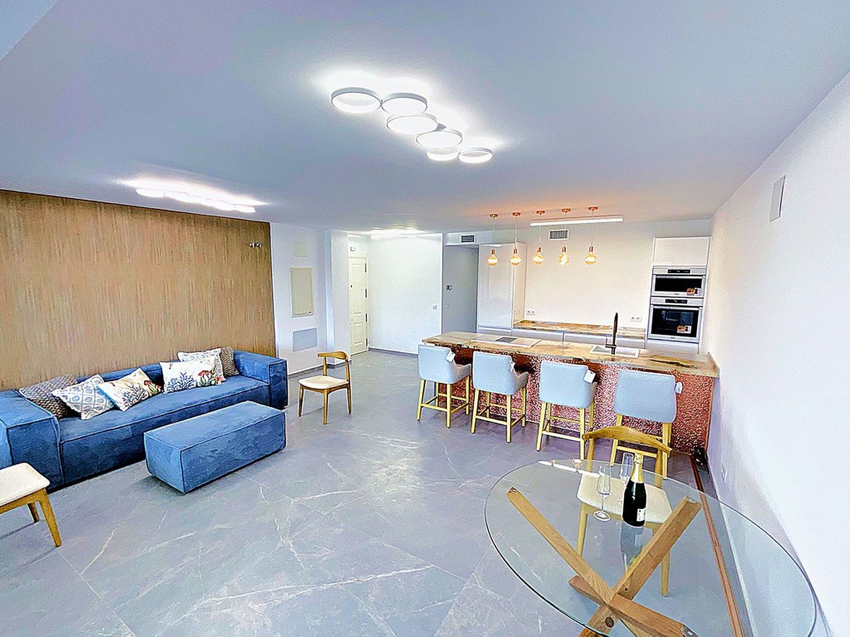 Modern open-plan living room with blue sofa, kitchen island, and glass dining table.