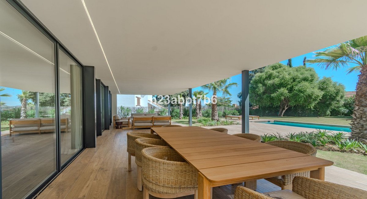 Modern covered patio with wooden dining table and chairs, overlooking a swimming pool and lush garden.