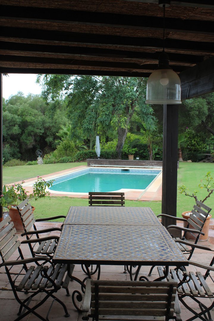 Covered patio with outdoor dining set overlooking a swimming pool and lush green garden.