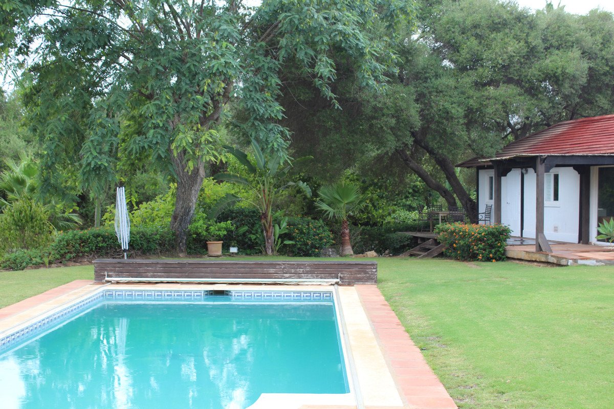 A swimming pool and a small cabin with a red roof set in a lush green garden with mature trees.
