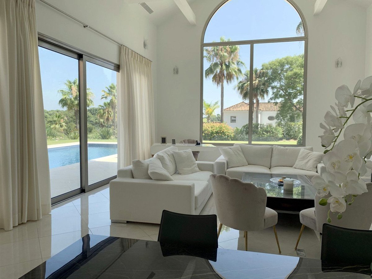 Bright living room with white sofas, large arched window, and sliding glass doors overlooking a pool and palm trees.
