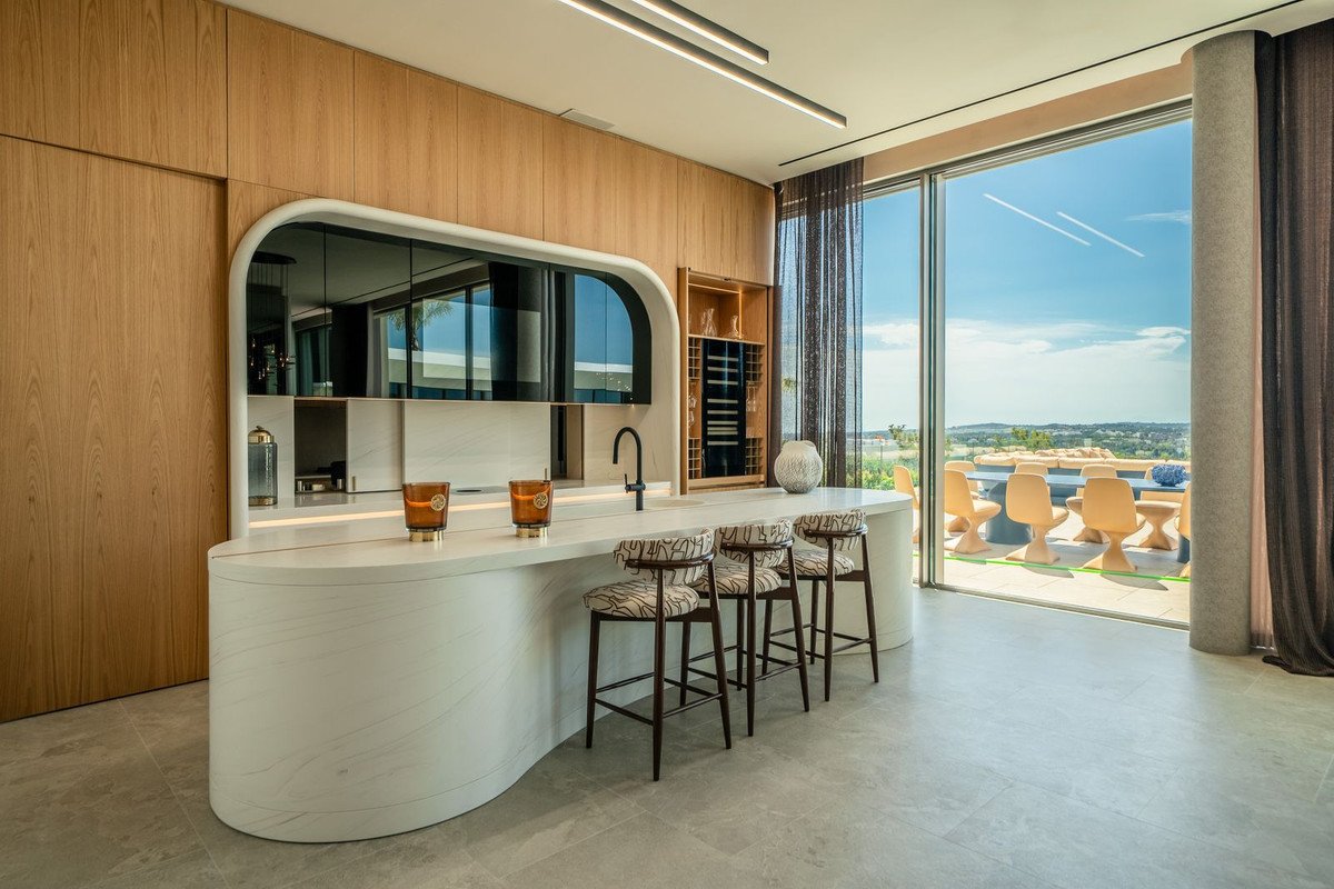 Modern kitchen island with bar stools, overlooking a patio with dining furniture and a scenic landscape view.