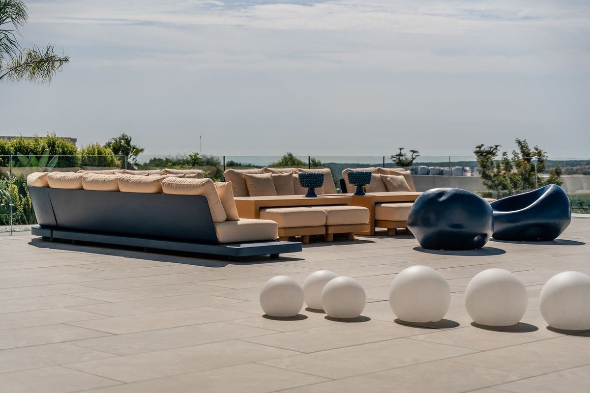 Modern outdoor lounge seating area with comfortable cushions, unique sculptural chairs, and a view of the sea under a clear sky.