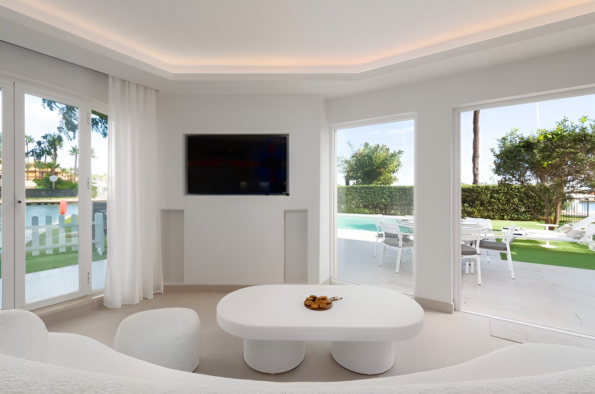 Modern white living room with a curved sofa, coffee table, TV, and sliding doors opening to a pool and outdoor seating area.