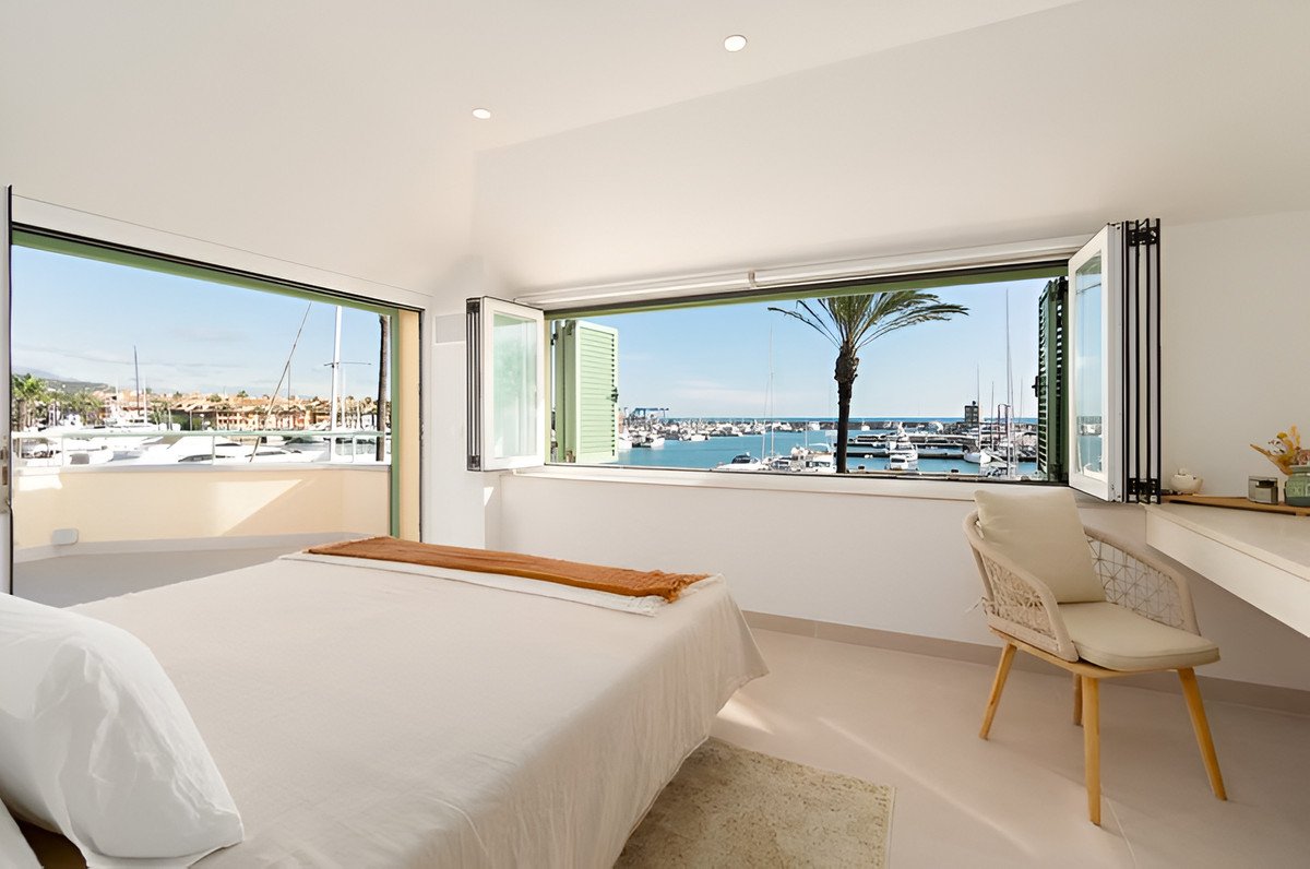 Bedroom with a large window offering a view of a marina, boats, and a palm tree. Features a desk and chair.