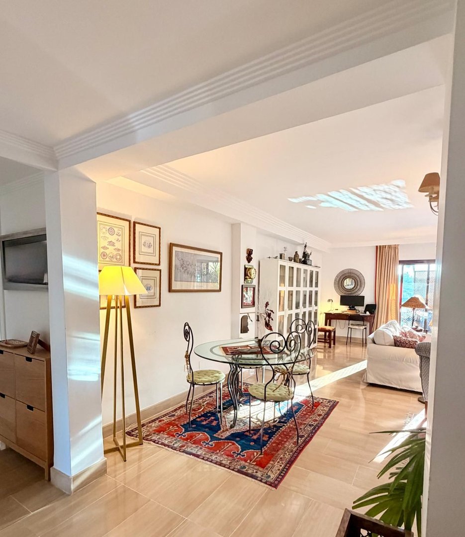 Bright and airy dining and living area with a glass table, chairs, sofa, desk, and natural light streaming in.