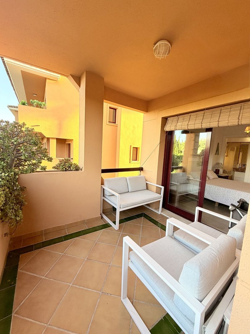 A furnished terrace with modern seating, overlooking a bedroom through glass doors, with a view of external architecture.