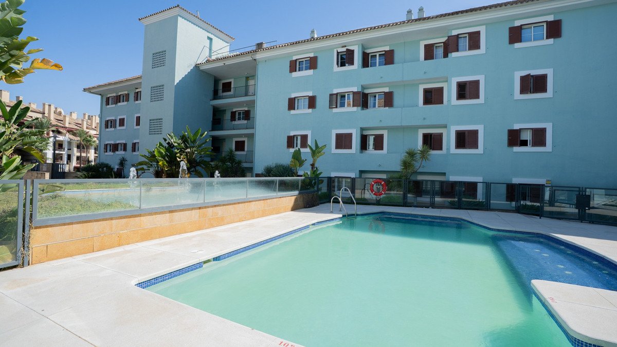 A turquoise swimming pool with a shallow end is in front of a light blue apartment building with brown shutters. Lush greenery surrounds the pool area.