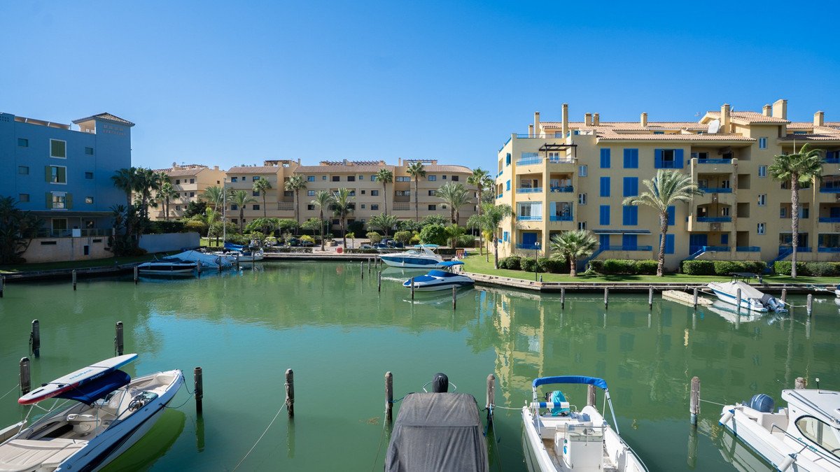 Apartment buildings overlooking a marina with boats docked, palm trees, and a clear blue sky.