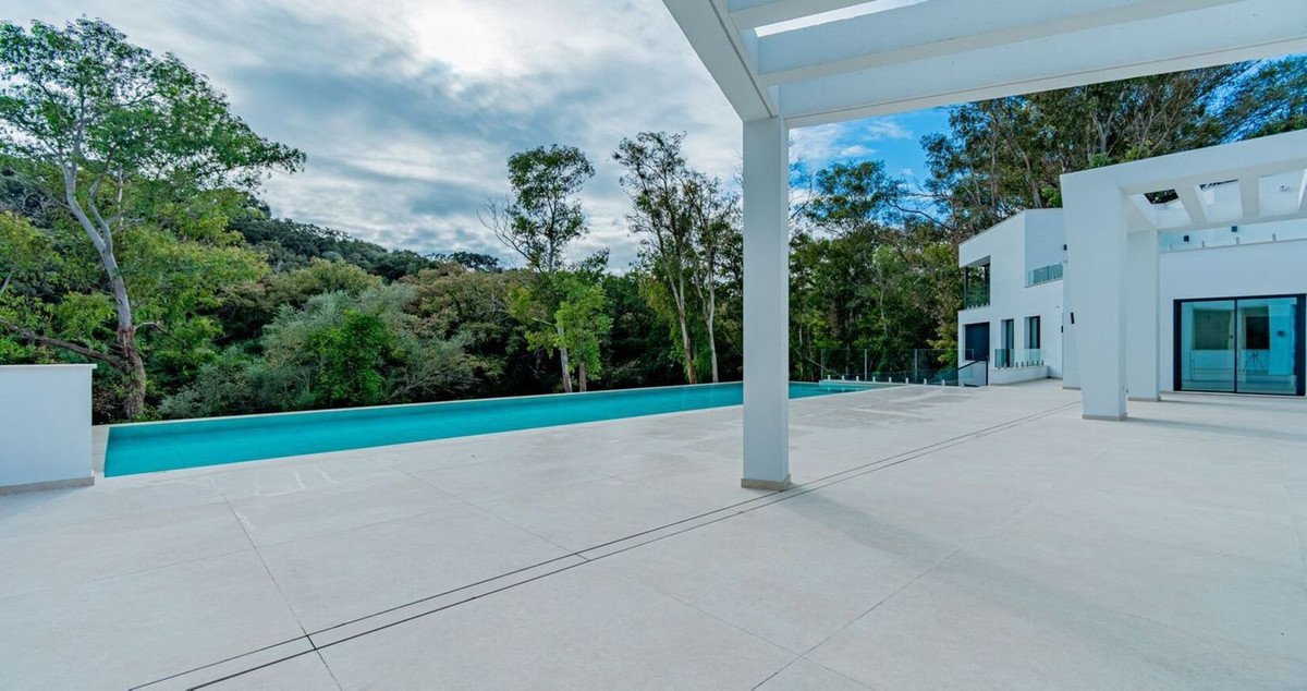 A modern white villa with an infinity pool and a lush green forest backdrop under a cloudy sky.