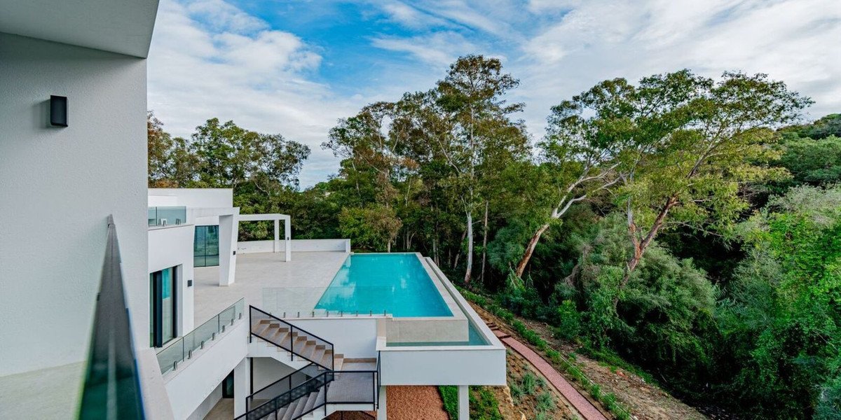 Modern villa with an infinity pool overlooking a lush forest under a partly cloudy sky.