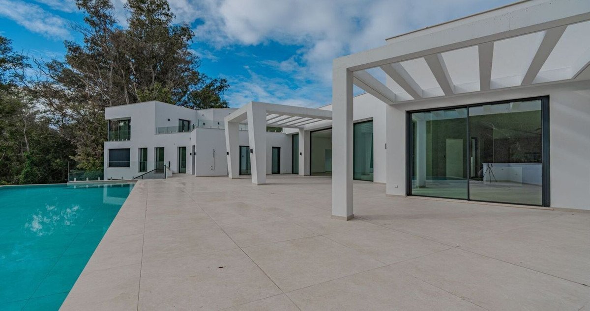 Modern white villa with a large swimming pool and spacious outdoor terrace, surrounded by trees under a blue sky with clouds.
