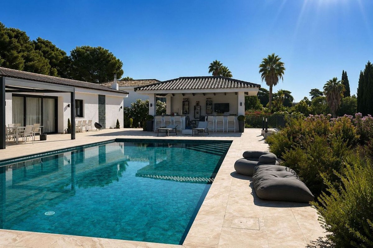 Luxurious outdoor swimming pool with a bar and lounge area, surrounded by lush greenery and palm trees under a clear blue sky.