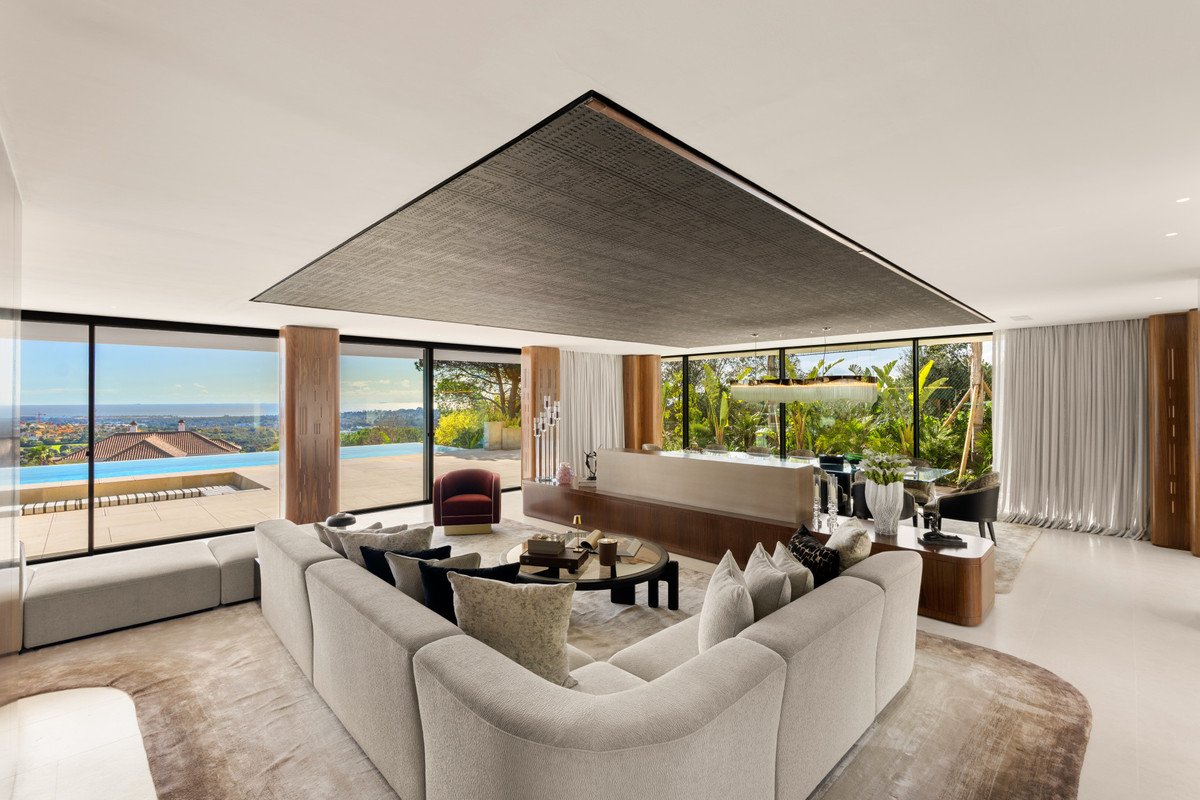 Spacious modern living room with large sectional sofa, panoramic windows, infinity pool, and lush garden views.