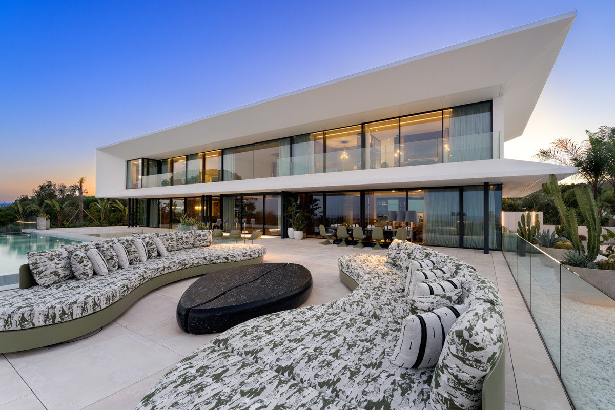 Luxurious modern villa with expansive terrace, curved sofas, pool, and desert-style landscaping at sunset.
