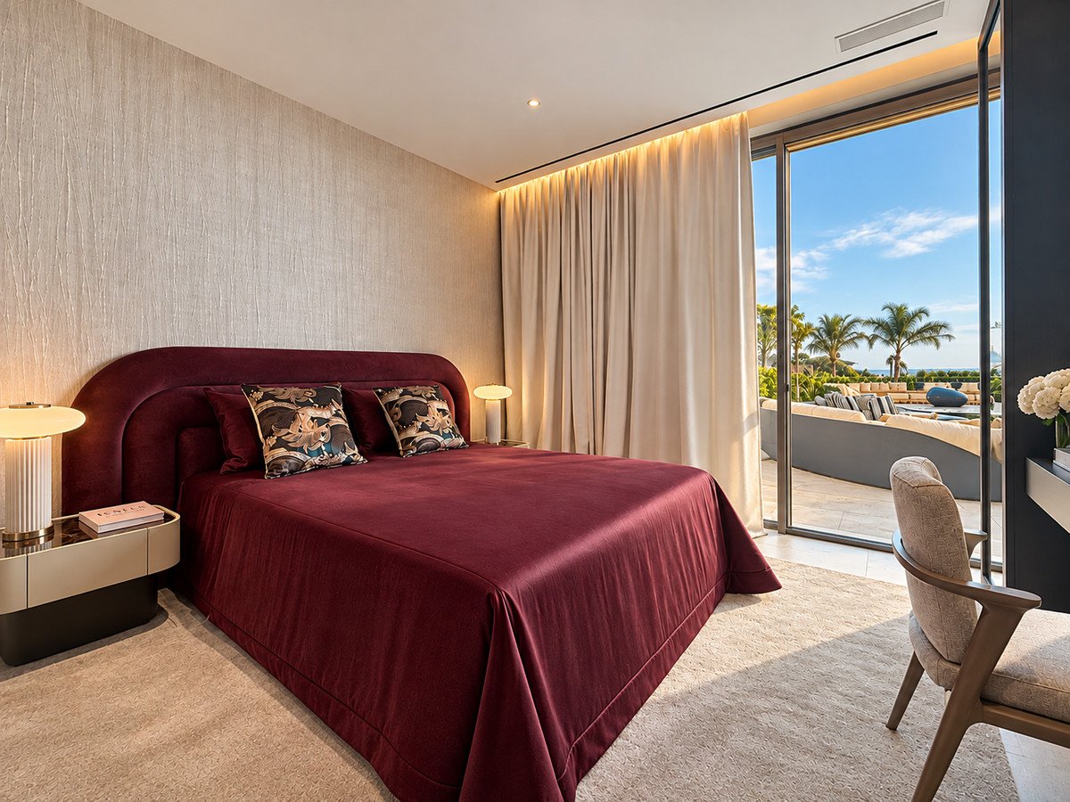A luxurious bedroom with a maroon bed, patterned pillows, and glass doors opening to a terrace with sea views and palm trees.