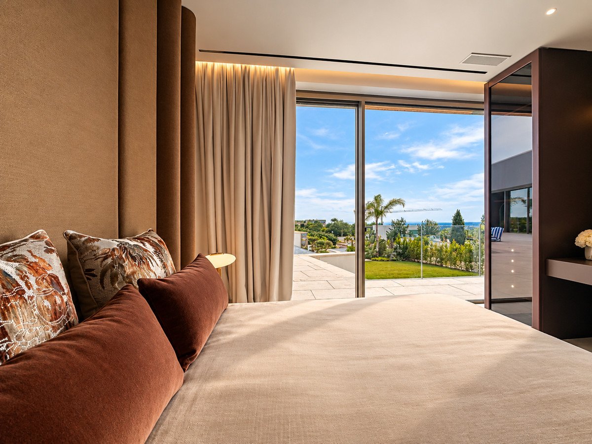 A bedroom with a large bed, large windows, and a view of the ocean and a green garden with a terrace.