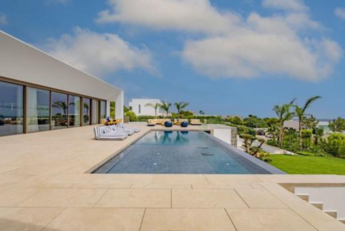 Modern villa with an infinity pool and a spacious terrace overlooking lush greenery and the distant sea under a blue sky.