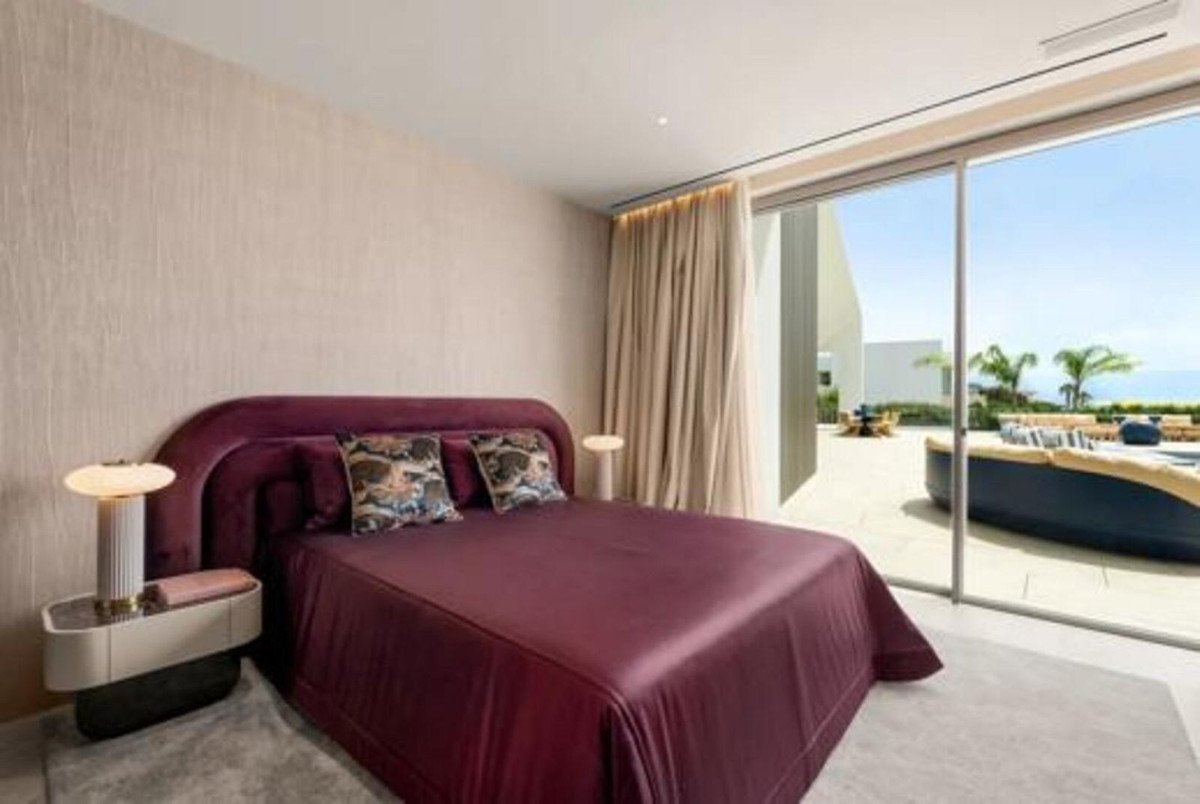 Modern bedroom with a large bed, decorative pillows, and a sliding glass door revealing a patio with a sea view and outdoor furniture.