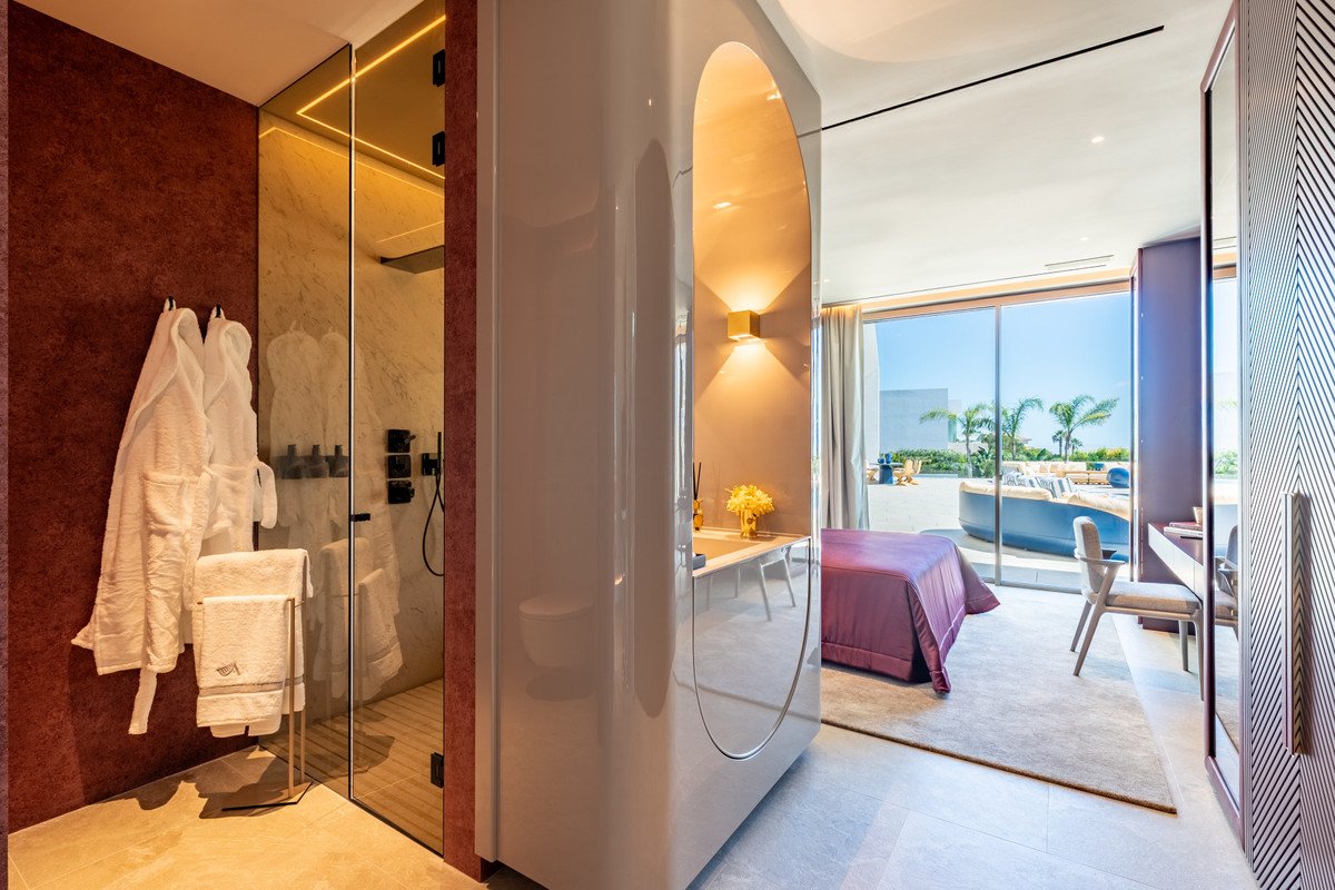 Modern bedroom with a purple bed, ensuite glass shower, and large windows overlooking an outdoor seating area and ocean view.