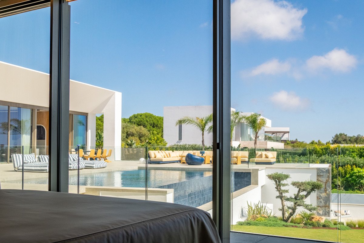View from a bedroom through a glass door to a modern villa with a swimming pool, terrace, lounge chairs, and lush green surroundings.