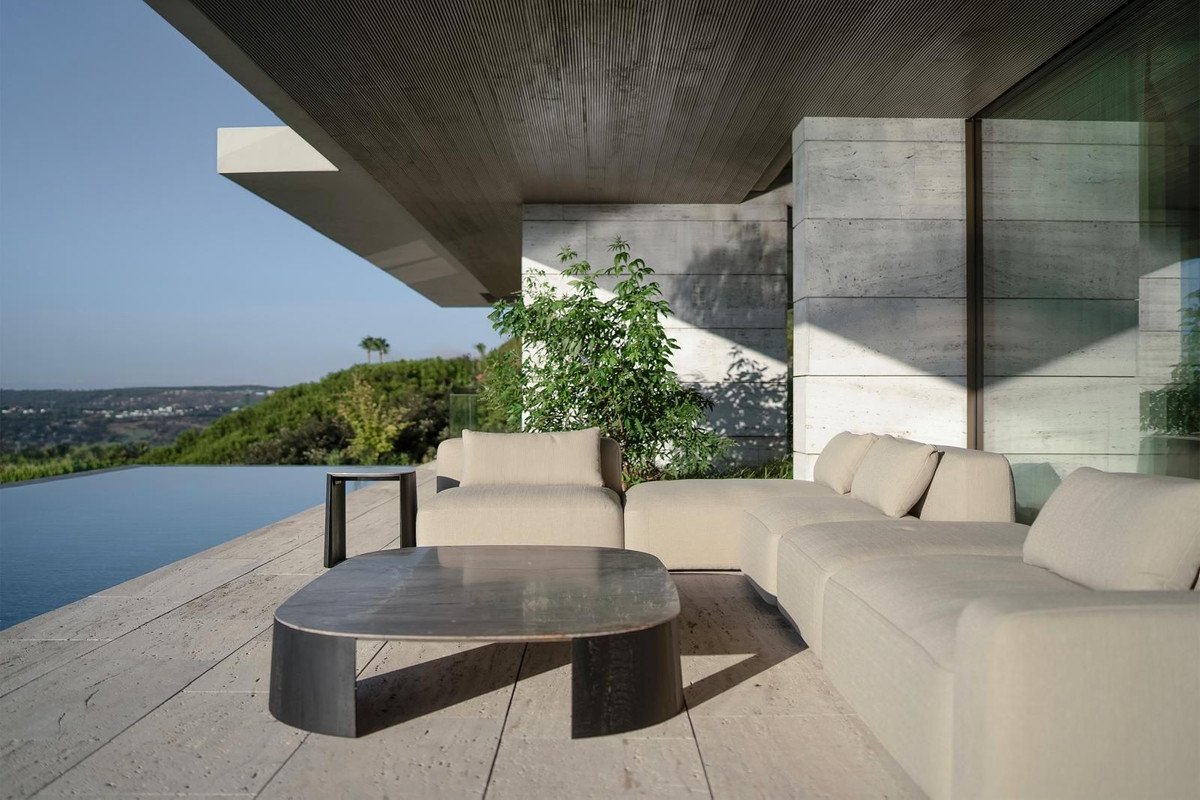 Modern patio with beige sectional sofa, coffee table, and infinity pool overlooking a lush green hillside and distant town.