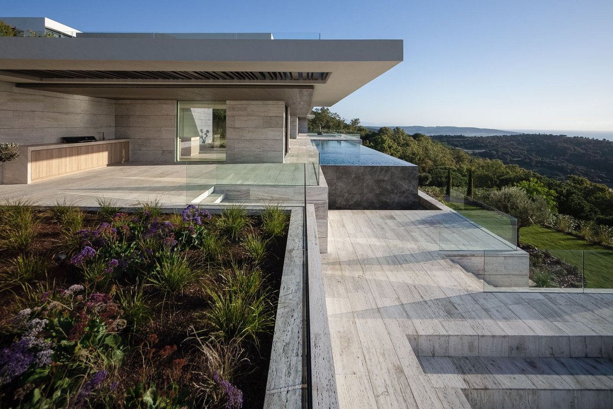 Modern villa with infinity pool and outdoor kitchen, overlooking a lush green landscape with rolling hills and a distant sea.
