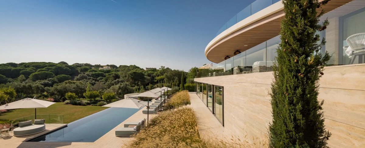 Modern villa with an infinity pool, lounge chairs, and lush green trees under a clear blue sky.