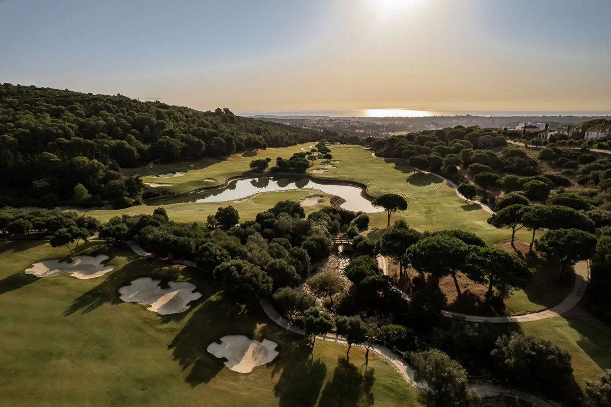 An aerial view of a picturesque golf course with a winding lake, bunkers, lush green fairways, trees, and a distant sea view.
