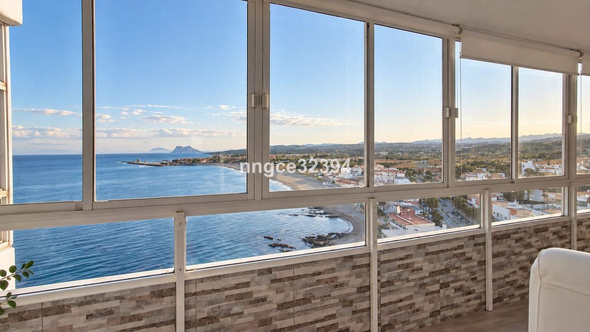 View from a balcony with large windows overlooking the sea, beach, and town with mountains in the distance.