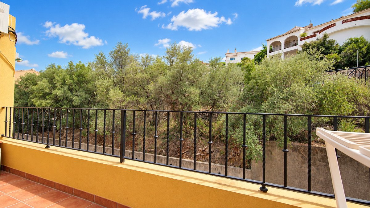 Balcony view with green trees and a bright blue sky with clouds. Houses visible on the hill.