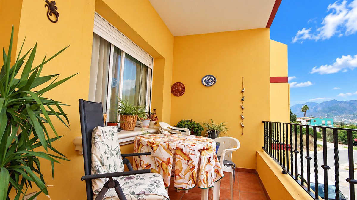 A yellow balcony with a table, chairs, plants, and a view of mountains and the town below.