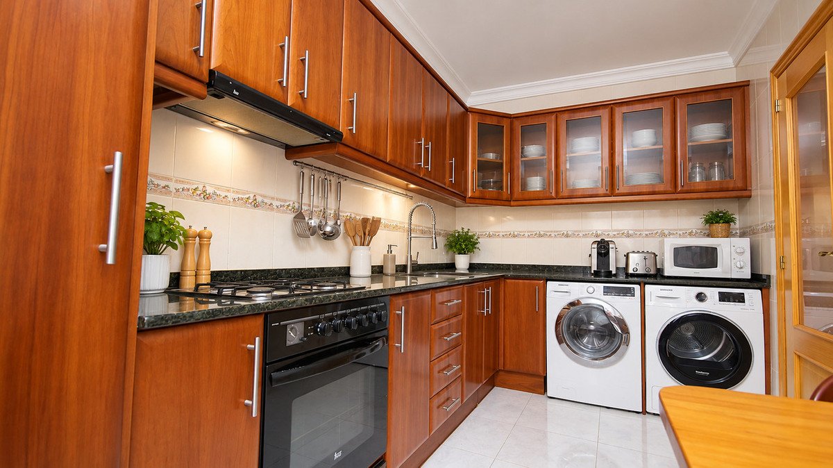 A modern kitchen with warm wood cabinets, featuring a stove, sink, microwave, coffee maker, and washing machine.