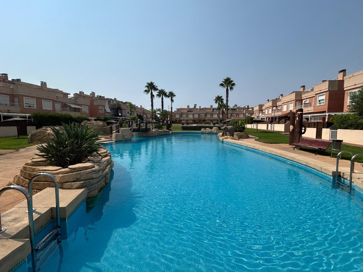 A large blue swimming pool with steps and a ladder, surrounded by green grass and townhouses with palm trees.